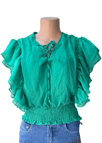 Vista de frente. Una blusa verde con volantes y detalles fruncidos en la cintura, y un lazo en el escote.